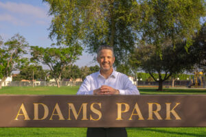 Person smiles while standing behind an "Adams Park" sign with hands resting on the top rail, surrounded by trees, open grass, and a playground in the background.