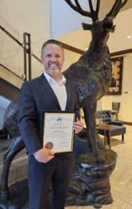Phil Bacerra smiles while holding an Elks "Elk of the Year" award certificate beside a large elk statue inside a lodge.