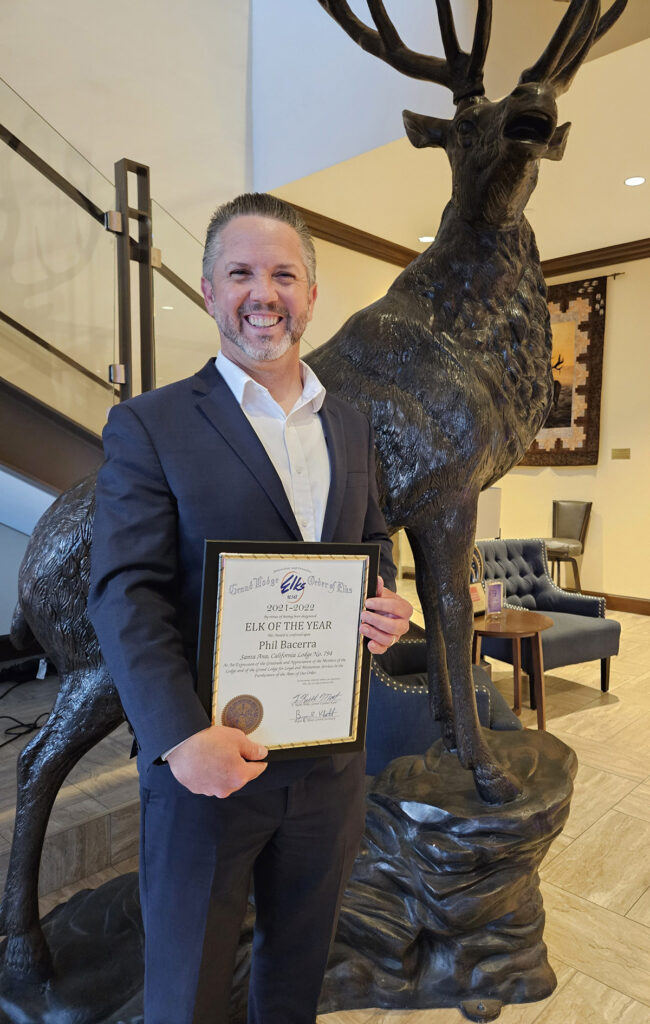 Phil Bacerra smiles while holding an Elks "Elk of the Year" award certificate beside a large elk statue inside a lodge.