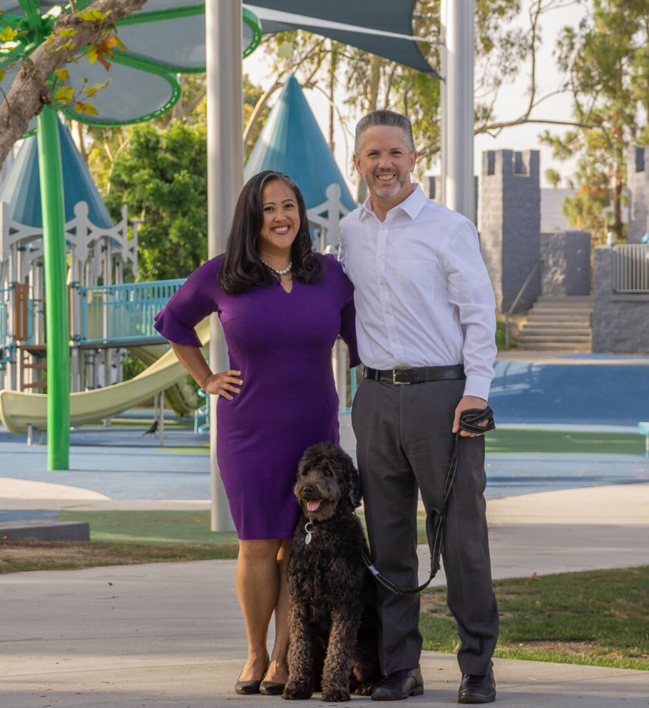 Phil Bacerra and his wife stand with their leashed dog on a park pathway in front of an inclusive playground featuring slides, climbing structures, shade canopies, and castle-themed play elements.