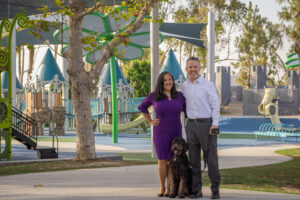 Two adults stand with a leashed dog on a park walkway in front of an accessible playground with slides, climbing structures, shade canopies, and trees.