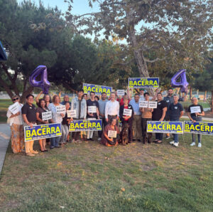 Community members gather in a park holding "Re-Elect Phil Bacerra" campaign signs and neighborhood name placards during a group campaign event for Santa Ana City Council Ward 4.