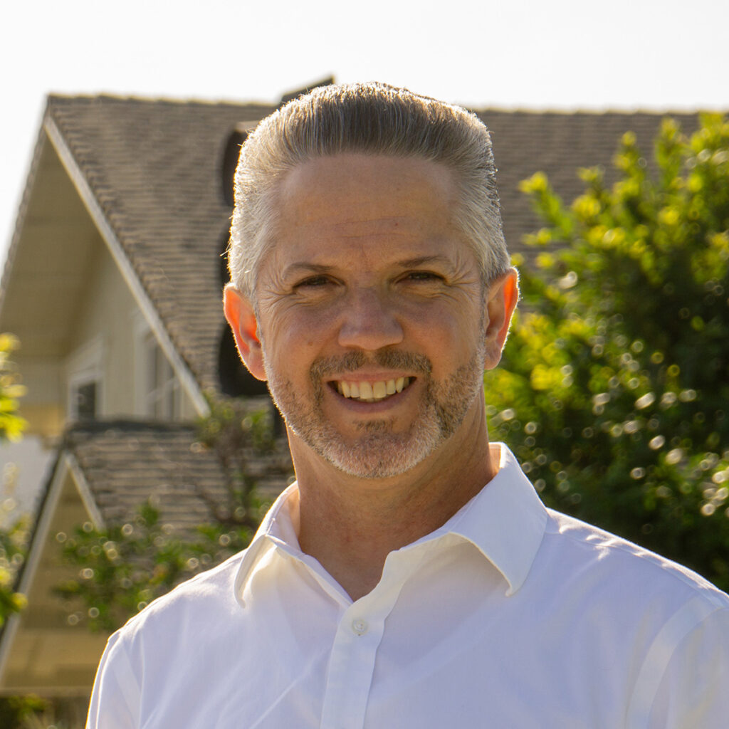 Phil Bacerra smiles outdoors in a white button-down shirt, with a residential home and trees softly blurred in the background.