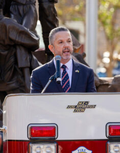 Phil Bacerra speaks at a podium during a public ceremony, with a microphone in front of him and a firefighter memorial sculpture in the background.