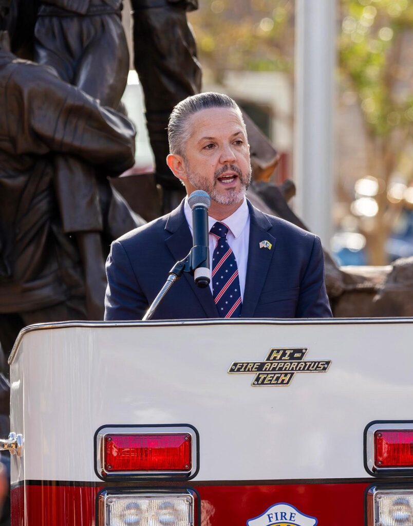 Phil Bacerra speaks at a podium during a public ceremony, with a microphone in front of him and a firefighter memorial sculpture in the background.